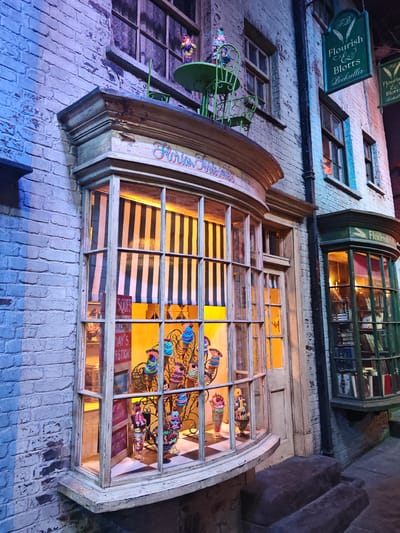 This is a photograph of a shopfront on Diagon Alley at the Warner Bros. Studio Tour London in Watford, United Kingdom. The building is constructed of painted brick. On the left is a large display window with black and white striped awnings, and a sign above that reads "Florian Fortescue's". Inside the window are various ice cream sundaes and signs for "Day's Questions" and "Bel Raisin". To the right is a green shopfront, and hanging above are green signs for "Flourish & Blotts Bookseller". A black pipe runs vertically between the two shopfronts. A green table and chair set is perched above the "Florian Fortescue's" window. A stone step leads to the shopfronts.