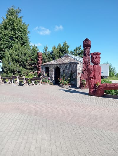 In Škilinpamūšis, Lithuania, on a clear, sunny day with a blue sky, a paved area with interlocking brickwork serves as a courtyard. To the left, several rustic wooden benches are arranged in a row, supported by stone bases. Behind the benches, a dense line of evergreen trees provides a natural backdrop. Dominating the center is a small, rustic structure with a stone facade and a metal roof, featuring an arched entrance. Flanking the structure and extending into the foreground are intricately carved wooden sculptures. One sculpture depicts a tall figure wearing a crown-like headdress, with another smaller figure beside it. To the right, a large, reddish-brown wooden sculpture of a person with a headdress and a visible beard is prominent, with a carved log extending from its right side. Smaller wooden figures are nestled amongst potted flowers, including red roses and pink petunias, that line the base of the stone building and surround some of the sculptures. In the background, beyond the trees, a white truck with a trailer is visible, suggesting the presence of a road or industrial area nearby. The scene appears to be an outdoor display or recreational area.