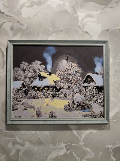 The image shows a framed oil painting of a snowy village scene, possibly located in the village of Okhotnichye, Russia. The painting depicts several rustic houses with snow-covered roofs and chimneys emitting plumes of smoke. Trees laden with snow surround the houses, and a dimly lit path or clearing is visible in the foreground. The overall atmosphere is serene and cold, typical of a winter landscape. The painting is signed "19.01.20".