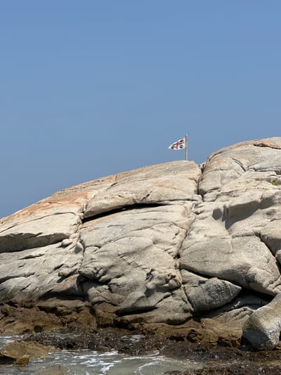 A flag with a white background and red cross is attached to a pole on top of a rocky outcrop. The flag is partially obscured by the rock. The outcrop is made of large, grey, weathered rocks with some patches of orange-brown discoloration. The rock is positioned above the sea, with a small area of water visible in the foreground. The sky is clear and blue. The scene is likely located on the coast of Castiadas, Italy.