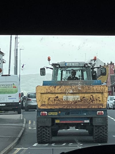 The image shows a close-up, slightly angled view of a large yellow and blue tractor, likely a snow plow given the weather. The tractor is on a road, which appears wet and possibly icy. Snowflakes are visible in the air, indicating it is currently snowing or has recently snowed. The scene suggests a cold, overcast day.
The tractor has a large yellow hopper at the back, likely for carrying salt or snow. Its front features a snowplow attachment. A license plate on the front of the tractor reads "132-DL-2550". The driver's cabin is visible, with a person seated inside. Other vehicles are partially visible behind the tractor, including a white van with "PREMIER" and "LANDSCAPES" written on its side, suggesting a possible service vehicle. The road has white lines indicating lanes.
The overall setting appears to be a rural or semi-urban area, with the presence of the tractor suggesting road maintenance in response to adverse weather conditions. The limited visibility due to the snow and overcast sky contribute to a somber, functional atmosphere. The image seems to be taken from inside another vehicle, as indicated by the black border on the left and the perspective. The location is identified as Falcarragh, Ireland.