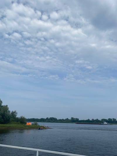 This is a landscape photograph of a wide river under a cloudy sky. In the foreground, a white railing suggests the viewpoint is from a boat or pier. The river occupies the lower half of the image, its surface showing ripples and reflections. On the left bank, a small, sandy area has an orange tent set up, with several people and two inflatable boats near the shore. The left bank is lined with dense green trees and foliage. Across the river, the right bank is also covered with trees. In the mid-ground of the river, a tugboat is pulling a long, flat barge. The sky is filled with a pattern of white and gray clouds against a pale blue background. The scene appears to be a natural setting near a body of water, possibly a recreational or camping area, and the specific location identified is Гольяны, Russia.