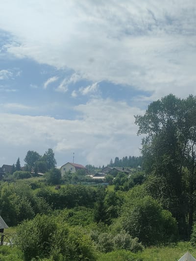 The image depicts a rural landscape on a partly cloudy day, with a village nestled amongst rolling hills covered in dense green vegetation and trees. Several houses with red and brown roofs are visible on the slopes. In the foreground, a large, leafy tree dominates the right side of the frame. The sky above is a mix of blue and white clouds, with some areas appearing brighter than others. The overall impression is one of a serene, natural setting, likely in a village or dacha community on the outskirts of Revda, Russia.