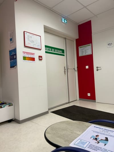 The image shows a hallway with a white emergency exit door marked "SORTIE DE SECOURS" in green. A red fire alarm is mounted next to a "violentomètre" poster. Below are smaller posters. A framed floor plan hangs on the wall. An exit sign is visible near the ceiling. Above the exit is a red wall section, a white door with restroom icons, and a document. A dark rug is on the floor next to a table with a sign.