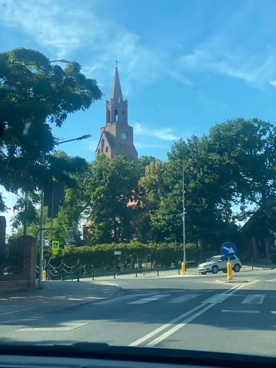 The image depicts a street scene in the city of Miłosław, Poland, dominated by a prominent brick church with a tall, pointed spire and a clock face. Lush green trees surround the church and line the streets, creating a verdant urban environment. A paved road with white crosswalk markings occupies the foreground, indicating a pedestrian crossing. Several streetlights are visible, along with traffic signs, including a blue circular sign with a white arrow indicating a mandatory turn. A light-colored SUV is parked on the side of the road, near a yellow bollard. A yellow pedestrian crossing sign with a silhouette of a person walking is also present. The sky is a clear blue with scattered white clouds, suggesting a pleasant daytime weather condition.