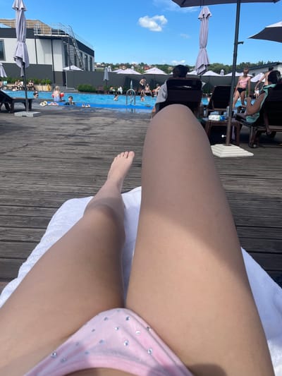 The image captures a sunny day at an outdoor swimming pool, possibly located in Oleshyn, Ukraine. The foreground is dominated by the legs of a person lying on a white towel on a wooden deck, wearing a pink bikini with scattered rhinestones. Beyond the deck is a bright blue swimming pool where several people are visible, including children and adults. Some are swimming, while others are relaxing by the poolside. Umbrellas provide shade around the pool area. In the background, a modern building with a black and white facade and a metal staircase stands prominently, suggesting a resort or a public facility. The sky is a clear blue with a few scattered clouds. The overall atmosphere is one of leisure and recreation.