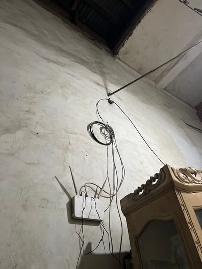 The image shows a white concrete wall inside a building in Kecamatan Waru, Indonesia. A white wireless router is mounted on the wall, with several black and white wires coming from it. A dark grey metal rod is attached to the wall above the router, with a cable running from it. There is a wooden cabinet to the right of the router, with a glass door and ornate carving on the top. The light is dim, illuminating the scene from the right side of the image. The photo appears to be taken at an upward angle, with the camera pointed slightly to the left.