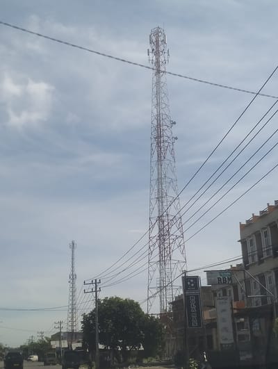 This image, captured in Mee Tanjong Usi, Indonesia, depicts a street scene dominated by a tall telecommunications tower in the background. The tower is a lattice structure, painted white and red, and is equipped with multiple antennae. In the foreground, there are several power lines strung across the frame. To the right, a multi-story building with a light-colored facade and visible windows is partially visible. Several signs are attached to the building, including one that reads "Kubota RBM." A smaller telecommunications tower is also visible in the distance on the left. The street is paved, and there are a few vehicles, including a black car and a white van, visible on the road. Trees and vegetation are present on the side of the road. The sky is partly cloudy, with a pale blue hue.