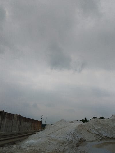 The image shows a large pile of white sand, likely limestone, in the foreground. The sand appears to have been recently moved as the surface is still wet and there are fresh tracks on the ground.  The pile is situated in front of a concrete wall with a small section of a red brick wall.  The wall runs parallel to the ground and is likely part of a larger structure. The background shows a cloudy overcast sky. The coordinates (latitude: 22.694, longitude: 90.601) place the location in the city of Pangasia, Bangladesh, suggesting this may be a construction site.  No people are visible.