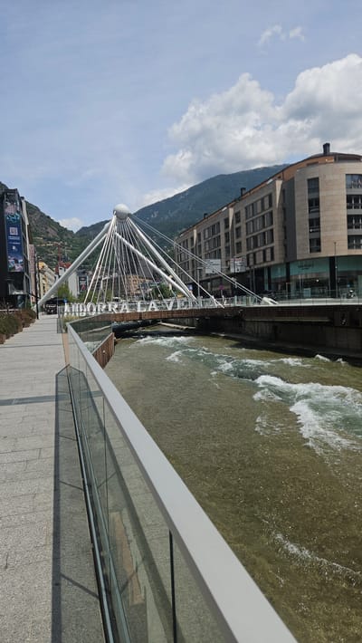 The image captures a detailed perspective of a city scene in Andorra la Vella, Andorra, under a bright, partly cloudy sky. The vantage point is from a paved stone walkway along the left bank of a river, looking towards a distinctive pedestrian bridge and the urban landscape beyond.
In the immediate foreground on the left, a light grey handrail with clear glass panels separates the granite-tiled walkway from the river. Shadows from the railing and an unseen object are cast on the walkway. The river, likely the Gran Valira, flows from the upper left towards the lower right, displaying visible rapids and white water indicating a moderate current; its water is a murky greenish-brown.
The central feature is a modern pedestrian bridge spanning the river. It is characterized by a prominent white spherical pylon in its center, from which numerous white suspension cables radiate downwards to support the brown-colored bridge deck. Large, white, capitalized letters spelling "ANDORRA LA VELLA" are affixed to the side of the bridge structure, facing the viewer. Several pedestrians are visible walking across the bridge.
Beyond the bridge on the left, multi-story commercial buildings with modern facades line the street, displaying storefronts and promotional banners. Trees and vegetation are visible on the lower slopes of the mountains behind these structures. On the right side of the river, across from the walkway, a row of contemporary multi-story buildings with large windows and light-colored facades (beige/tan) are visible. One building at a corner features a rounded architectural element. A prominent "MANGO OUTLET" sign is visible on a ground-level storefront, and various road signs are present further down the street.
The background is dominated by steep, tree-covered mountains that rise directly behind the city structures. The sky above is a light blue with scattered white, cumulus clouds. The scene is illuminated by strong daylight, creating clear shadows.