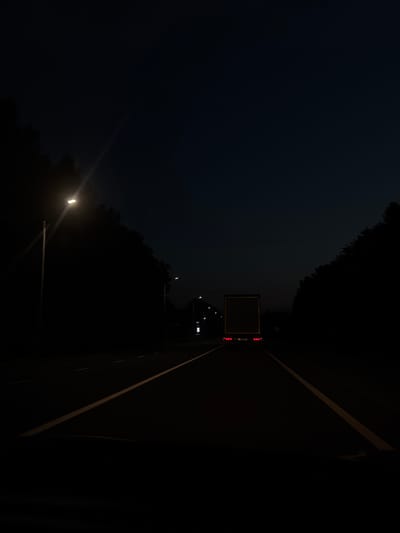 The image depicts a dark, rural highway at dusk or night, likely near Vargany, Russia, given the provided context. A large, box-shaped truck is the primary subject, illuminated by its own taillights and potentially ambient light from distant sources. The truck is traveling away from the viewer, positioned in the center of the roadway. Yellow lane markers are visible on the asphalt, dividing the road. To the left of the road, a streetlamp casts a bright light upwards and outwards, with its beam creating a visible streak against the dark sky. Trees and foliage line both sides of the highway, appearing as dark silhouettes against the faint light. The overall atmosphere is somber and quiet, with the absence of other vehicles or pedestrians. The sky is a deep, dark blue, suggesting either early morning or late evening twilight.