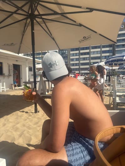 The image depicts a sunny beach scene in Knokke-Heist, Belgium. A shirtless man in patterned swim trunks sits on the sand, holding a glass cocktail garnished with fruit and mint. He wears a grey Under Armour baseball cap. Behind him are beachgoers, lounge chairs, and beach umbrellas, with tall buildings in the background. A large beige beach umbrella labeled "Schweppes" is visible to the left. The sand is light brown. The man is focused on his drink.
