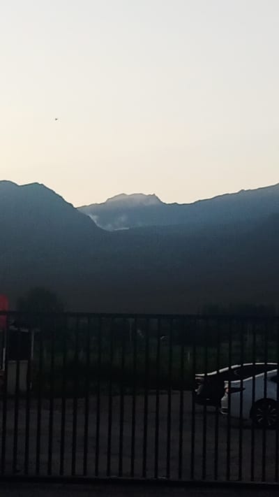 The image shows a wide view of a mountainous landscape at dusk or dawn. The sky is a pale, washed-out color, with a faint light suggesting the sun is low on the horizon. In the distance, dark, silhouetted mountains rise against the sky. A wisp of smoke or cloud is visible near the peak of one of the mountains, hinting at a possible natural phenomenon or human activity. In the foreground, a black metal fence runs horizontally across the frame, obscuring the lower portion of the scene. Through the fence, a dark, indistinct area suggests vegetation or perhaps buildings. On the right side, partially visible behind the fence, is the rear of a white car, its shape suggesting a modern hatchback or sedan. A small, dark object, possibly an airplane, is visible in the upper left quadrant of the sky. The scene appears to be located in a rural or semi-rural area, with the mountains serving as a prominent natural backdrop. Given the prompt, this scene is to be located in Choktal, Kyrgyzstan.