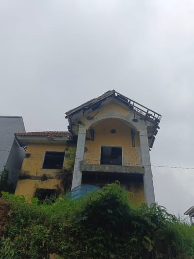 The image displays a two-story, yellow-painted, derelict building situated on an overgrown embankment in Makilala, Philippines, viewed against an overcast sky. The structure features a prominent arched entrance on its upper level, supported by two light-colored columns. The roof is significantly damaged with missing tiles and exposed framing. All visible window openings are empty. The yellow facade is faded, stained, and exhibits signs of graffiti or defacement. Dense green vegetation, including grass and ferns, covers the embankment in the foreground and extends onto the lower portions of the building. A partially visible blue cylindrical object, possibly a water tank, is nestled within this foliage. To the immediate left, a portion of an unfinished gray concrete structure is visible. A single black utility line traverses horizontally across the mid-ground. No individuals or dynamic actions are present in the scene.