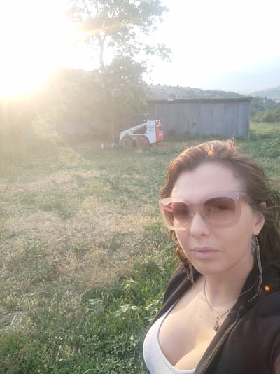 An outdoor, eye-level, waist-up selfie is captured in Spathades, Greece, under bright sunlight. A woman with long, brown hair, wearing large, square, light brown sunglasses, a grey tank top, and a black jacket, is the primary subject. She is adorned with a silver pendant necklace and long, dangly silver earrings.
The background features a grassy field with scattered dry patches and some green vegetation. To the left, a large tree casts a shadow, and bright sunlight flares into the lens. In the mid-ground, a white and red Bobcat skid-steer loader is parked near a corrugated metal shed. Beyond the shed, rolling hills covered in greenery and trees recede into the distance, under a hazy sky. The overall scene suggests a rural or semi-rural setting during daylight.