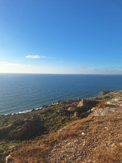 The image depicts a coastal landscape under a clear blue sky with a few scattered white clouds. The foreground is dominated by dry, brown vegetation and rocky terrain. A white plastic bag is visible on the ground in the mid-foreground. A steep, rocky coastline covered with sparse green vegetation slopes down towards the ocean. The sea is a deep blue, with gentle waves lapping at the shore. The horizon is clear, with a faint outline of land visible in the distance. The overall scene suggests a natural, somewhat rugged coastal environment. The mention of Ahrara, Morocco, places this scene within a specific geographical context.