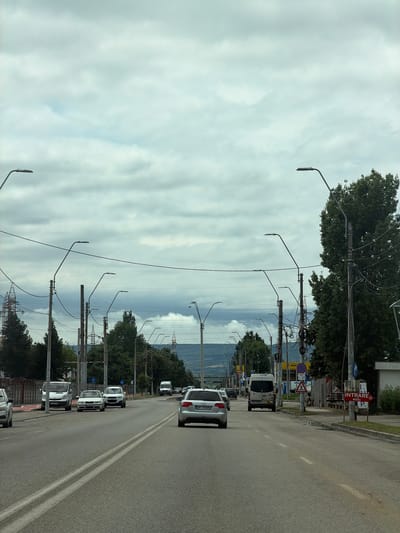 This media file captures a street scene in Drobeta-Turnu Severin, Romania, likely during daytime under an overcast sky. The image is taken from a slightly elevated perspective, looking down a two-lane road.
**Subjects:** The primary subjects include a long stretch of asphalt road with double solid white lines down the center, numerous utility poles fitted with streetlights and dense overhead wires, and a variety of vehicles. These vehicles consist of several white cars (sedans and what appears to be a van), some parked along the right side of the road, and others potentially in traffic. Lush green trees line both sides of the street, and in the distance, hills or mountains are visible, interspersed with large power pylons. The sky above is largely covered by diffuse clouds.
**Activity or event taking place:** The scene depicts typical daily activity on a street, with vehicles parked or moving.
**Notable details:** The weather is cloudy, contributing to a soft, even light across the scene. The utility poles are particularly prominent, with a multitude of wires crisscrossing overhead. On the right side of the road, several signs are visible. A red rectangular sign clearly reads "INTRARE P." (Romanian for "Entrance Parking"). Below it, a blue circular "no parking" sign is visible, along with a yellow sign that partially shows the text "PITII". A triangular warning sign for a pedestrian crossing is also present. The license plate "MH 99 MUN" is clearly visible on a white sedan in the foreground, and another plate starting "MH 02" is on a car further down, confirming the location in Mehedinți County, where Drobeta-Turnu Severin is situated. The overall impression is that of an active urban or suburban street.
