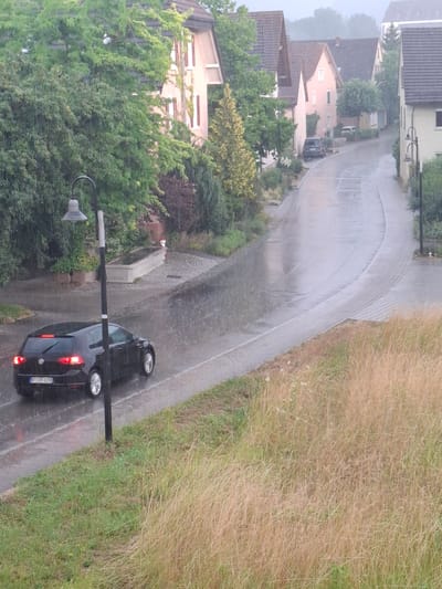 A black hatchback car, possibly a Volkswagen Golf, is driving on a wet asphalt road during a rain shower. The car's brake lights are illuminated, indicating it is slowing down or stopped. A street lamp is positioned to the left of the car, and a narrow sidewalk runs alongside the road. In the background, there are several houses with tiled roofs and trees lining the street. The overall environment suggests a residential area in Klettgau, Germany, experiencing inclement weather. The road surface is reflective due to the rain, and water is visible on the ground and falling from the sky. The surrounding vegetation appears lush and green.