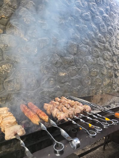 The image shows meat cooking on skewers over coals. The meat appears to be pork, with some pieces on skewers resembling kebabs and others appearing to be larger cuts of meat. Two sausages are also visible on skewers, and they are a reddish-brown color. The meat is positioned over a grate which is situated above a bed of glowing coals, indicating a cooking process. Smoke is rising from the cooking meat and dissipating into the air. The background consists of a stone wall, which has a textured, irregular pattern. The scene appears to be outdoors, possibly in a rural setting given the stone wall and the open-air cooking setup. The lighting suggests it is daytime, but the specific time is not discernible. The overall impression is one of food preparation, likely for a meal or gathering. The location context provided is Pidhorodne, Ukraine. There is no visible text in the image.
