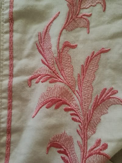 A close-up, high-angle shot of intricate pink embroidery on a cream-colored fabric, likely a garment or textile. The embroidery features stylized floral or leaf-like patterns with a prominent vertical border on the left side, also in pink embroidery. The fabric appears to be cotton or a similar natural material, with visible weave and slight creasing indicating it is not perfectly taut. The lighting is soft and diffused, highlighting the texture and detail of the embroidery. There are no people or specific landmarks visible in the image. The location in Daşoguz, Turkmenistan, is not discernible from the visual information provided in the image itself.