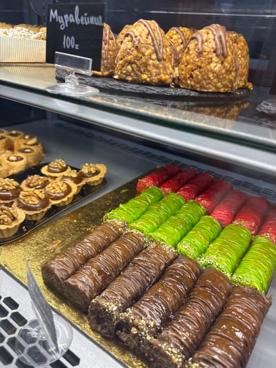 This is a close-up shot of a display case filled with various sweets, likely in a bakery or confectionery shop. The camera angle is angled down, focusing on the array of desserts.

In the foreground on the left, there are several stacked cookies that appear to be made with oats and nuts, drizzled with chocolate. A black sign with white lettering is attached to the display, showing Cyrillic text and a price: "Муравейник 100г". This translates to "Ant Hill 100g", suggesting it's a type of pastry named for its appearance.

To the right, a selection of rolled desserts is arranged. These appear to be baklava or similar sweets, with layers of dough rolled into cylinders. They are presented in three different colors: red, bright green, and brown. The red and green ones are sprinkled with what looks like crushed pistachios or nuts. The brown ones are coated in a glossy chocolate glaze and also appear to have nuts sprinkled on them. These are arranged in neat rows on a golden display tray.

Further down and to the right, another tray holds smaller, individual tarts. These tarts have a fluted edge and are filled with a dark, smooth topping, likely chocolate or a similar sweet paste, garnished with walnut halves.

The setting appears to be indoors, suggested by the artificial lighting and the glass display case. There are no people visible in the frame. The overall impression is of a well-stocked and visually appealing display of sweet treats. The location context provided is Alakhadzi, Abkhazia.