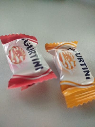 Two individually wrapped candies are on a light gray, textured surface. The candy on the left is wrapped in red and white foil, and the candy on the right is wrapped in orange and white foil. Both candies have "GURTINI" written in black capital letters on their wrappers. The candies appear to be lying on a countertop in a room in Krolevets, Ukraine.