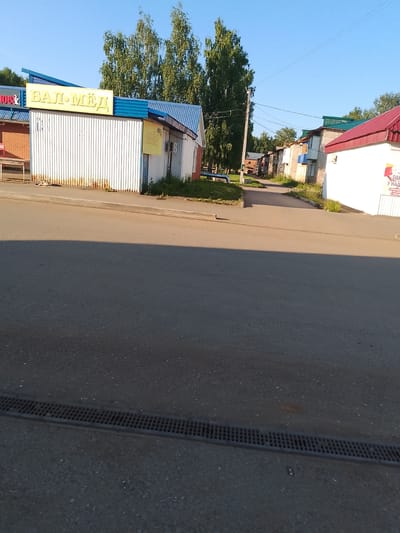 The image captures a street scene in Kranokholmskiy, Russia, under a clear blue sky. A building with a white corrugated metal facade and a blue roof is prominently featured on the left. A yellow sign with Cyrillic text "БАЛ-МЕД" (BAL-MED) is affixed to the building. To the right of this building, another structure with a white facade and a red roof is visible. A poster with Cyrillic text, including "ОДА У НАД..." (ODA U NAD...), is attached to its wall. In the background, several residential buildings with brick facades and various colored roofs, as well as tall trees, line the street. The foreground consists of an asphalt road with a black drainage grate running across it. The scene appears to be a commercial or residential area on a sunny day.