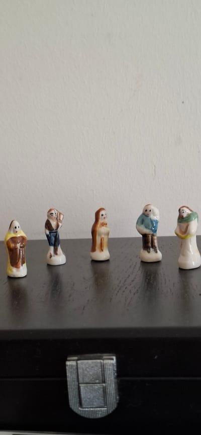 The image shows five small, ceramic figurines standing in a line on a dark, reflective surface against a textured, off-white wall. The figurines appear to be religious figures, possibly monks or nuns, dressed in robes of various colors including brown, yellow, white, and blue. They are all facing forward, with subtle differences in their posture and attire. The background is a plain wall with some faint texture. The dark surface they are standing on might be a piece of furniture, possibly a box or a shelf, with a metallic latch visible at the bottom of the frame. The lighting is soft and diffused, casting gentle shadows. There is no indication of the city of Le Havre, France, or any other specific location.