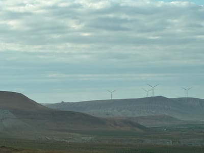 The image depicts a wide, somewhat desolate landscape under a cloudy sky. In the foreground, rolling hills and scrubland stretch across the frame, with some texture suggesting sparse vegetation or dry earth. In the mid-ground and background, larger, more rugged hills rise, their slopes marked by erosion and geological layers.

Prominently featured on the crests of these distant hills are several wind turbines. Four are clearly visible, silhouetted against the sky. They are modern, three-bladed wind turbines, indicating a renewable energy installation. One wind turbine is isolated on a hilltop further to the left, and another group of three is visible on a ridge to the right. A faint, man-made structure, possibly a building or ruin, is also visible on a distant ridge to the far left.

The sky is filled with a thick layer of overcast clouds, creating a diffused, soft light. There are hints of blue sky peeking through in some areas, suggesting a break in the clouds or a transition in the weather. The overall impression is one of a vast, open, and somewhat muted natural environment, punctuated by modern energy infrastructure. The time of day appears to be daytime, given the overall brightness, but the cloud cover makes it difficult to determine precisely. The location context of Gobustan, Azerbaijan, is consistent with such a landscape featuring wind farms.