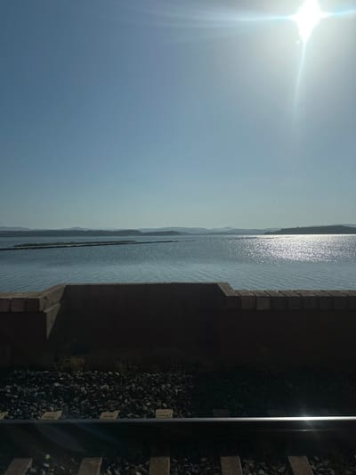The image shows a bright, sunny day at a body of water. In the foreground, there is a brick wall and train tracks. Beyond the wall and tracks is a large expanse of water, with a long, thin structure or island in the middle of it. In the distance, there are hills and mountains. The sun is very bright and high in the sky, causing a glare on the water. The location is the Narbonne region of France, which is known for its coastal areas and lagoons.