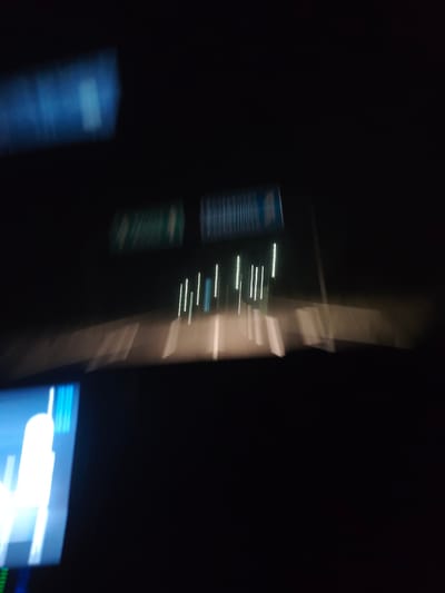 An out-of-focus, nighttime scene taken from inside a moving vehicle. The vehicle is traveling on a highway or well-lit road. Road signs are visible, but illegible due to the blur. There are white lines on the road and streetlights or reflectors on the right side of the road. Inside the vehicle, a dashboard screen is visible on the bottom left of the frame and another blurry object appears at the top left. The image is too blurry to provide further details. There is no evidence that this image is in Barakovo, Bulgaria.