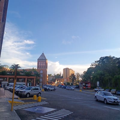 The image depicts a parking lot outside a commercial building in Carrizal, Venezuela. The architecture of the building features a prominent brick clock tower and a domed structure. The parking lot is occupied by several vehicles, including SUVs, sedans, and a white pickup truck. Pedestrian crosswalks and accessibility markings are visible on the asphalt. The sky is blue with scattered clouds, suggesting it is daytime. Trees and other buildings are visible in the background, indicating an urban or suburban environment. The overall scene is a typical view of a commercial area with adequate parking facilities.