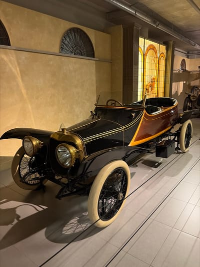 An antique or vintage car is displayed indoors, likely within a museum or exhibition space in Wassenaar, Netherlands. The vehicle is predominantly black with a polished wooden upper body or trim, accented with gold-colored pinstripes. It features distinctive brass components, including two sets of headlights and other fittings, and rests on white-walled tires with black wire-spoked wheels.
The car is positioned on a light-colored tiled floor, casting a long, distinct shadow on the adjacent wall, which is also tiled. In the background, a large, ornate stained-glass window with intricate patterns in warm amber and orange hues suggests a historic or grand architectural setting. Above the window, industrial-style pipes run along the ceiling. Further into the background, yellowish walls feature decorative circular wrought-iron grilles. The lighting indicates an indoor, artificially lit environment, creating a focused and atmospheric presentation of the automobile. No people are visible in the image, and no clearly legible text is present on the car or in the surroundings.
