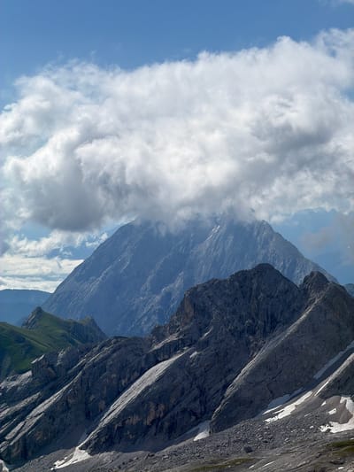 The image depicts a mountainous landscape under a partly cloudy sky. In the foreground, rugged, rocky terrain slopes upwards, with patches of snow visible in the crevices. A series of dark, jagged mountain ridges dominates the middle ground, with some areas covered in shadows. In the background, a large, imposing mountain peak rises, partially obscured by a thick blanket of white clouds. Further in the distance, the outlines of other, more distant mountains are faintly visible against the blue sky. The scene is devoid of any human presence or man-made structures, suggesting a remote natural environment. This is a view from the area of Wildermieming, Austria, showcasing the alpine topography.