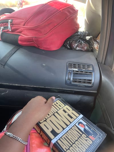 The image depicts the interior of a vehicle, observed during daytime in Hoss, Nigeria. A dark red fabric backpack, appearing somewhat worn, rests on the vehicle's dark-colored dashboard on the left. Adjacent to it on the right, a black plastic bag, tied at the top, is also on the dashboard. Further to the right on the dashboard, an air conditioning vent with horizontal slats and a circular control knob is visible, exhibiting minor cosmetic damage with white scuff marks. Below the vent, an "AIRBAG" label is present. In the lower foreground, a person's right hand with dark skin and a silver-colored chain-link bracelet holds a copy of the book "Unlimited Power" by Anthony Robbins, which is labeled a "NATIONAL BESTSELLER." Through the vehicle's window in the upper left background, blurred bright outdoor light and indistinct shapes, including a red object resembling a vehicle or motorcycle, are visible.