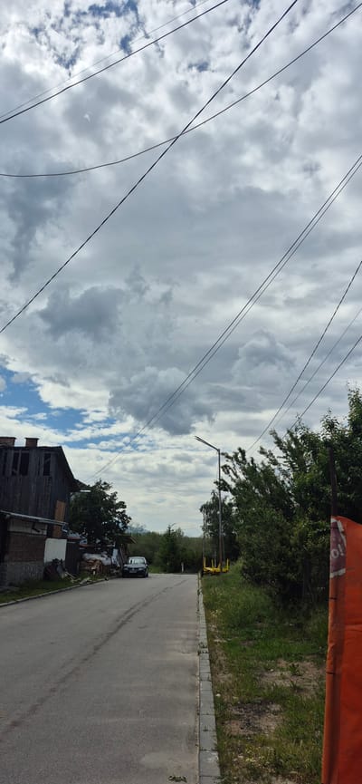 The image presents a daytime scene of a street in a semi-rural setting, captured under a predominantly cloudy sky with patches of blue. The photograph appears to have been taken with a vertical orientation, but its content is best viewed horizontally.

In the upper portion of the frame, several dark, thin utility lines crisscross the sky, which is filled with an expanse of grey and white clouds.

The lower half of the image is dominated by an asphalt road that stretches into the distance, showing signs of wear, dirt, and faint tire marks. A concrete or stone curb borders the right edge of the road, separating it from a grassy area and dense green trees and bushes that line the path. A tall utility pole with an extended arm, possibly a streetlight, is visible further down the road on the right side. In the distance, a small white vehicle is partially obscured by vegetation.

On the left side of the road, a dilapidated, two-story house with a weathered wooden upper section and a brick foundation is visible. Piles of wood and debris are scattered near the house. A dark-colored car, possibly a sedan, is parked on the shoulder of the road facing away from the viewer.

In the immediate foreground, intruding into the upper right corner of the original image (when correctly oriented, it would be in the foreground to the right), is a large, out-of-focus orange object, which could be part of a barrier or construction equipment. Faint, reddish text, possibly "TO" or part of a longer word, is vaguely visible on its surface but is illegible.

The scene conveys a quiet atmosphere, devoid of human activity, and the architecture hints at an older, possibly Eastern European, locality consistent with Dobrinishte, Bulgaria.