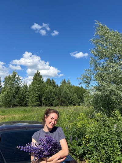 A young woman with brown hair pulled back stands outdoors, leaning against the rear of a dark-colored car. She is holding a large bouquet of purple flowers. The woman is wearing a grey t-shirt and smiling at the camera. The background is a landscape with a field of green grass and wildflowers, a dense forest of evergreen and deciduous trees, and a bright blue sky with scattered white clouds. The location is identified as Verkh-Rozhdestvo, Russia.