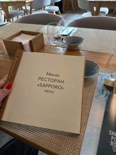 A menu for the "Sapporo" restaurant, with text in Russian, rests on a woven placemat on a wooden table. Glasses and chairs are also visible. There is no information to determine this image was taken in Peresvet, Russia.