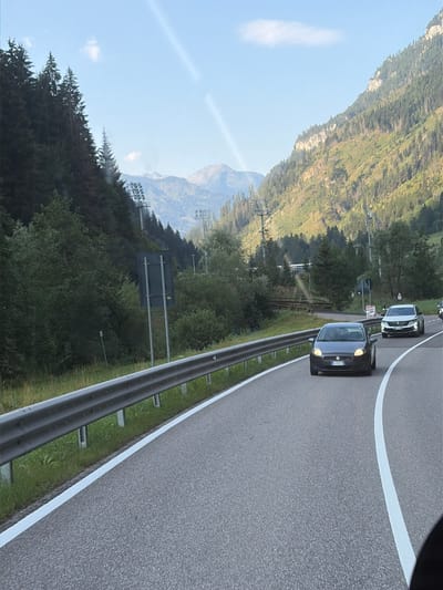 The image depicts a scenic mountain road in Predazzo, Italy, likely captured from within a vehicle. In the foreground, a dark-colored Fiat car is traveling on the right side of the asphalt road, which is marked with white lines. A metal guardrail runs along the left side of the road, separating it from a grassy embankment and a dense forest.
The surrounding environment is characterized by a steep, wooded mountainside on the left and a sun-drenched, rocky mountain slope on the right. The sky is clear blue with a few wispy clouds. In the distance, a series of pylons, possibly related to ski lifts or power lines, are visible ascending the mountainside. The lighting suggests it is daytime, with the sun casting strong shadows and illuminating the mountainous terrain. The overall impression is one of a picturesque alpine landscape.