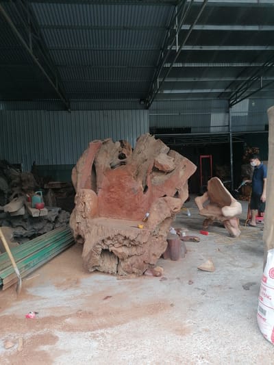 The image captures the interior of a workshop or warehouse in Ha Tu Ward, Vietnam. The space is filled with wooden creations, primarily what appear to be chairs crafted from large, irregular pieces of wood. One chair, significantly larger and more prominent, features a rough, raw texture with a reddish hue. Another chair, smaller and smoother, sits nearby. A man wearing a mask is seen in the background, possibly working on or inspecting one of the wooden pieces. Tools, wood scraps, and a bag of material are scattered on the concrete floor, indicating ongoing or recent woodworking activity. The building has a corrugated metal roof and walls, contributing to a utilitarian atmosphere.
