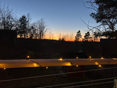The image shows a twilight scene, likely in Gränsta, Sweden, given the prompt's information. A wooden railing with small, warm-toned string lights runs horizontally across the lower third of the frame. Beyond the railing is a yard with dormant grass. In the middle ground, partially obscured by the railing and lights, is a small, red structure, possibly a shed or outbuilding. The background consists of a dark silhouette of trees against a gradient sky ranging from dark blue at the top to orange near the horizon, indicating a sunset or sunrise. There are no people visible in the image. The scene suggests a rural or suburban environment. The lighting is dim, with the string lights providing the primary source of illumination.