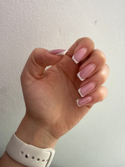 A close-up image depicts a human left hand, oriented with the palm facing towards the viewer and fingers slightly flexed. The hand displays a light skin tone. All five fingernails are present, featuring a French manicure style with a translucent pink base and opaque white tips. The nail shape is squoval. A white silicone smartwatch band is visible on the wrist, secured with a metallic clasp pin inserted into one of the strap holes. The background is a plain, light-colored, textured wall. The scene is located in Senokos, Bulgaria.