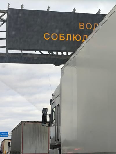 The image depicts a traffic scene, likely on a highway in Dorokhovo, Russia, based on the road sign indicating direction towards Moscow. A large electronic sign is visible above the road, displaying a message in Russian, spelled out in yellow LED lights. The message reads "ВОД СОБЛЮД," which may be an abbreviation or partial word in the Russian language. A line of trucks is present on the road, with a large white truck occupying a significant portion of the right side of the frame. The sky is overcast, suggesting cloudy weather. The overall impression is one of a busy transit route with traffic flow control measures.