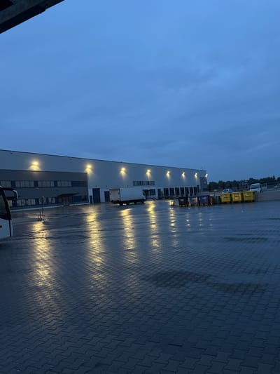 The image depicts an outdoor scene at dusk or early morning in Bielany Wrocławskie, Poland. A large, modern industrial building with numerous loading docks is visible. The building is illuminated by several overhead lights. In front of the building, a paved area, likely a parking lot or loading bay, is wet, indicating recent rain or damp conditions. Lights from the building reflect on the wet surface, creating streaks of illumination.
Several objects are present in the paved area:
* A white truck is parked to the right of the loading docks.
* A row of large, colored bins (black, blue, and yellow) are lined up further to the right.
* A white vehicle, possibly a van or small truck, is parked on an elevated ramp to the far right.
* Part of a white vehicle, possibly a bus or large van, is visible on the left side of the image, closest to the camera.
The sky is overcast and a deep blue, suggesting low light conditions. The overall impression is one of a functional, industrial area during a period of low visibility. There are no people clearly visible in the image. The environment is urban or industrial, with the building and pavement dominating the scene.