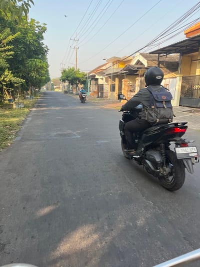 A medium shot captures a person riding a black motorcycle on a paved road in Karangtanjung, Indonesia. The rider, facing away from the camera, wears a black helmet and a gray jacket over dark pants, with a blue and gray backpack secured on their back. The motorcycle, identified by the license plate "W 3982 NEA" with an expiration date of "02-28," has a red taillight illuminated. The road is bordered by trees and grassy verges on the left, and houses with yellow and orange facades on the right. Utility poles with numerous wires are visible along the street. Another person on a motorcycle is seen further down the road in the distance. The sky is clear and light blue, suggesting daylight. The scene appears to be a residential street in a suburban or semi-urban area.