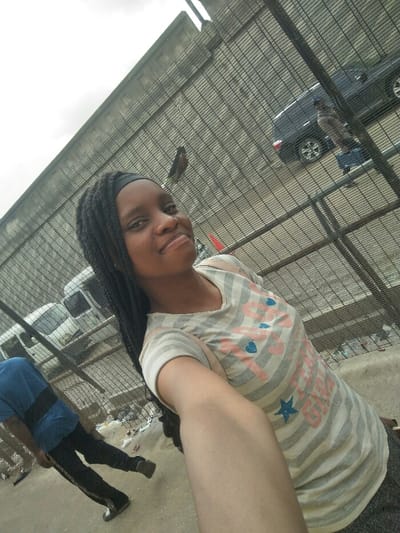 A young female, identified as the primary subject, is taking a selfie outdoors in Ajah, Nigeria. She is positioned in the foreground, slightly angled, extending her right arm towards the camera. She has a dark complexion, wears a black headband, and her long, dark braided hair falls over her left shoulder. She is dressed in a light-colored, possibly off-white or light grey, horizontally striped t-shirt with "1997" printed in pink, along with blue heart and star motifs. Her expression is a faint smile, with her eyes directed at the camera.
A dark metal grid fence separates the foreground from the background street scene. Through the fence, a paved road is visible, displaying varying amounts of litter and a small orange traffic cone near the center-right. On the road, a dark-colored SUV is visible to the right, and a white van is present to the left. Two individuals are also visible in the background street area: one male figure, partially obscured on the left, wearing a blue t-shirt with a horizontal black stripe and dark trousers, walking away from the camera. Another individual is faintly visible on the right, behind the dark SUV, wearing light-colored upper clothing and blue lower clothing. The sky is overcast, and a concrete structure is visible in the upper background, beyond the fence.