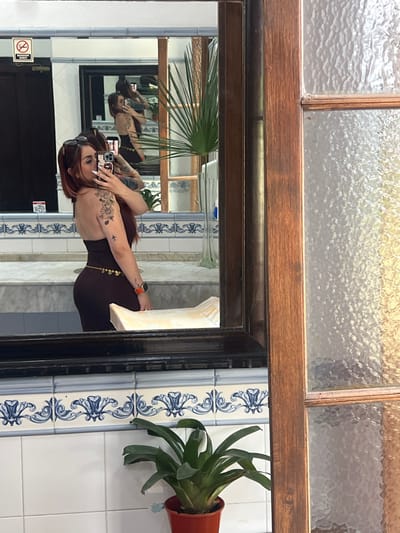 A woman is visible in a mirror, taking a selfie with her phone. She has red hair, tattoos on her arm, and is wearing a tight-fitting brown dress. The reflection shows her in a public restroom, with a white tiled wall, blue patterned tiles, and a black door. A plant in a brown pot is attached to the wall. Above the mirror is a window with textured glass and a dark wooden frame. Another mirror can be seen in the reflection.