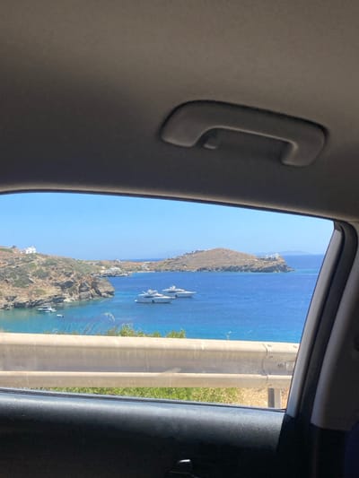The image depicts a bright daylight coastal scene in Φάρος, Greece, viewed from inside a vehicle. The foreground shows the vehicle's light gray headliner, a dark gray overhead grab handle, and the dark gray window frame and upper door panel. Outside, a light-colored two-bar metal guardrail extends horizontally across the mid-ground, indicating a roadside elevated above the sea. Sparse dry vegetation and green foliage are visible below the guardrail. The central view encompasses a deep blue bay with clear, lighter turquoise water in shallows. Two large white motor yachts are anchored in the bay's center-right, while at least two smaller white boats are present in a cove on the left. The bay is framed by arid, rocky hillsides covered with sparse scrub. Several white buildings, characteristic of the region, are situated on these hills, including a prominent structure on a distant promontory to the right and another on the hillside to the left. The sky is clear and bright blue. No individuals are visible; the scene is static.