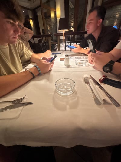 Two young men are seated at a table covered with a white tablecloth. One man in the foreground, wearing a tan shirt, has curly brown hair and is looking upwards and to his right. The other man, in the background, wears a black shirt and is looking down at his phone, which he holds in his right hand. He also appears to be wearing a black watch.

On the table, there are two glass ashtrays, cutlery (forks and knives), and what appear to be menus with QR codes. A red lighter with "VIKING CRUISES" printed on it is also visible. The scene is dimly lit, suggesting it is nighttime or indoors. The presence of menus and cutlery indicates a dining setting, possibly a restaurant. The atmosphere seems casual, with both individuals engaged with their phones. The man in the background is holding his phone in his left hand as well. The man in the foreground is wearing bracelets on his left wrist.

The provided location context is Rüdesheim, Germany.