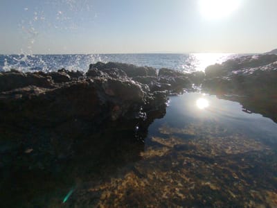 A low-angle, wide shot captures a rocky shoreline at what appears to be Karaburun, Turkey. The foreground features dark, wet rocks, one forming a natural pool that reflects the bright sun and the sky. The water in the pool is clear, revealing the textured seabed beneath. In the mid-ground, rough, dark rocks jut out towards the sea, with waves crashing against them, sending spray into the air. The sea stretches to the horizon, meeting a clear, pale blue sky. The sun is high and bright, creating a strong glare on the water's surface and a lens flare in the lower right portion of the image. The overall impression is of a rugged coastal landscape bathed in bright sunlight.